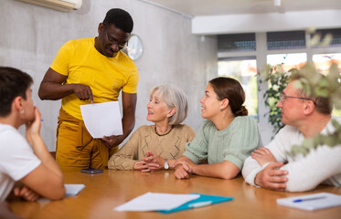 Fototapeta premium Interested adult African American man participating in volunteer work discussing project or event with group of associates, men and women of different ages and nationalities gathered around table
