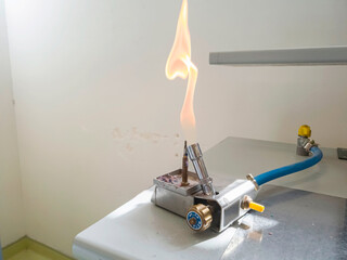 A Bunsen burner ignited with a large shaped, orange-colored flame on a laboratory bench. A blue gas supply pipe connects to a yellow gas stopcock. A regulating valve labeled "methane".