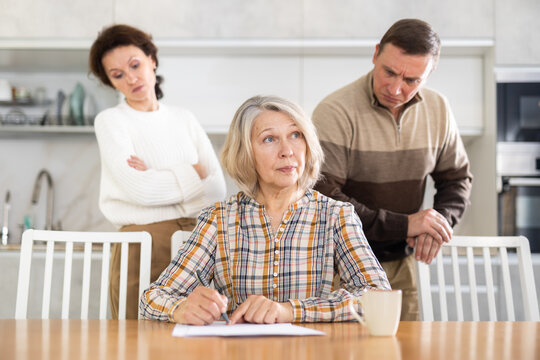 Thoughtful old woman compiling heritage document while middle-aged man and woman competing with each other in the kitchen
