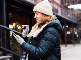 Fototapeta premium A woman in winter clothes and a hat holds a smartphone while typing a message in a winter city.