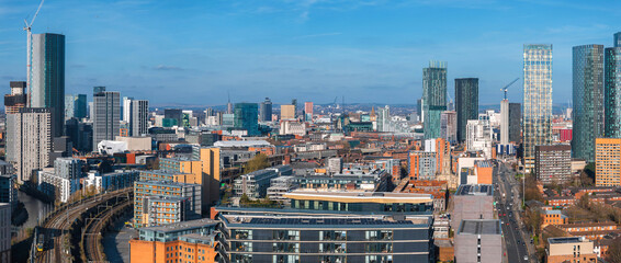 Aerial view of Manchester, UK, showcasing skyscrapers, construction cranes, a railway line, and a mix of modern and historic architecture under a clear sky.