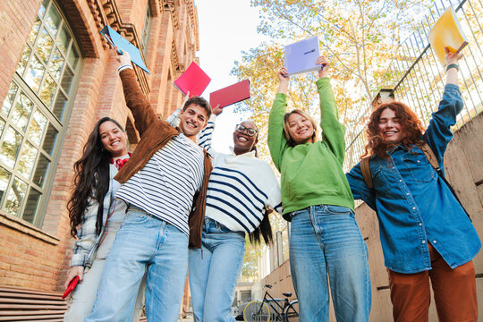 Group of enthusiastic students cheerfully lifting their notebooks in a triumphant academic success, embodying the spirit of teamwork and friendship during their time at school and university.