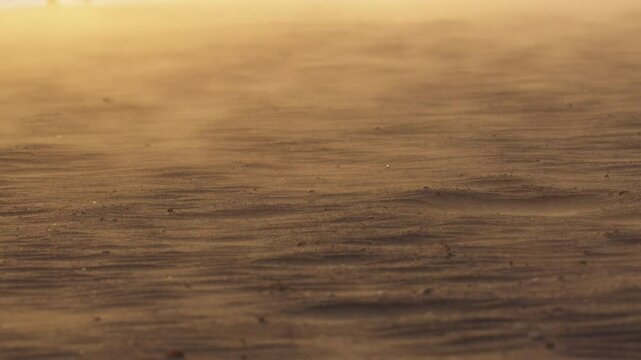 Golden sunset casting warm light over arid desert landscape, wind sweeping fine sand grains and creating dynamic, flowing patterns across desolate terrain