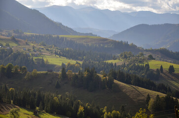 Obraz premium Picturesque view of lush green valleys, forested hills, and distant mountain ranges under a partly cloudy sky with sun rays shining through. Carpathian Mountains, Ukraine
