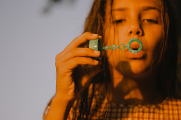 A young girl creates bubbles outdoors, focusing on the ephemeral beauty and joy of childhood activities. Soft, warm sunlight. Kid blowing bubbles.