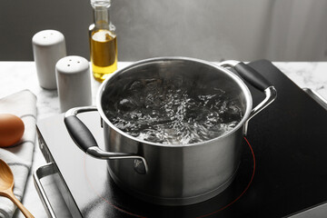 Cooking pot with boiling water and stove on light table against grey background, closeup