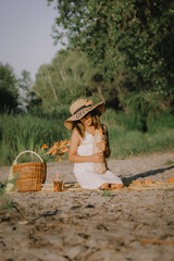 Obraz premium A girl wearing a wide-brimmed hat and white dress enjoys a picnic on a sandy bank, holding bread and looking down. A picnic basket. Summer vacation.