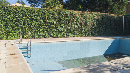Blue tiled walls and steps of an empty swimming pool. Background wall of green leaves. Vacation low...