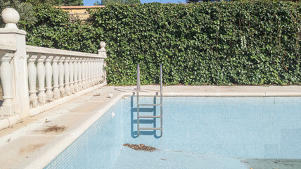 Blue tiled walls and steps of an empty dry swimming pool. Background wall of green leaves. Vacation low season. Square pattern of blue ceramic tiles, mosaic. Summer recreation in Spain
