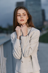 Fototapeta premium Young woman in light beige jacket posing outdoors at dusk with thoughtful expression, urban background and muted color palette, fashion and psychological concept