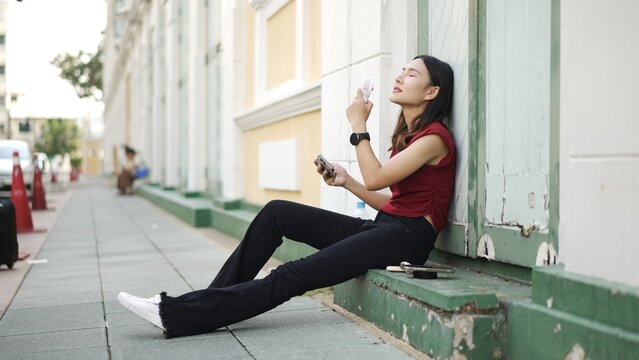 Young Asian woman sitting on city sidewalk, holding smartphone and enjoying cool breeze from portable fan, relaxing alone during hot summer day outdoors