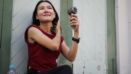 Young Asian woman sitting outdoors in urban area, using modern handheld fan while flipping her hair and smiling, enjoying cool breeze during hot summer day © pixs4u