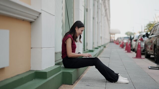 Young asian woman in casual outfit sitting on step and working on laptop in city street, surrounded by parked cars and cones, showing modern freelance urban lifestyle.