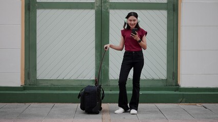 Young Asian woman traveling with suitcase, using smartphone while listening to music on wireless...