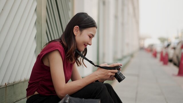 Young Asian female photographer sitting peacefully outdoors, smiling while reviewing photos on her camera, expressing creativity, freedom and quiet joy in modern lifestyle during travel.