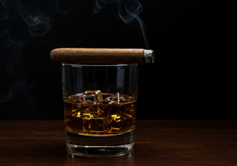 Glass of whiskey next to a lit cigar on a wooden table. Relaxing ambiance with warm lighting in the background, creating a cozy and inviting setting.