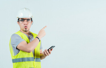 Surprised builder worker with phone pointing at blank space isolated. Amazed engineer holding phone...