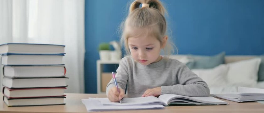 A focused blonde child writes in her workbook beside a tall stack of books on a tidy bedroom desk. Concept of early learning discipline and childhood education.

