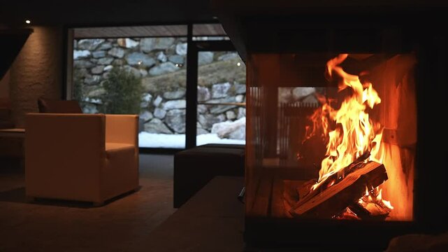 A modern fireplace with flickering flames in a cozy lodge interior. Large windows reveal a snowy alpine scene with a stone wall and sparse greenery.