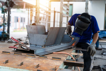 Fototapeta premium An industrial worker performing TIG welding on a curved metal structure in a factory workshop. The scene captures real work conditions with tools and equipment on the table.