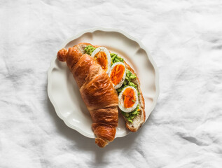 Croissant sandwich with avocado and boiled egg on a white ceramic plate on a light background, top view