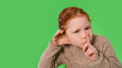 Young girl with red hair making a shushing gesture while wearing a cozy sweater against a bright...