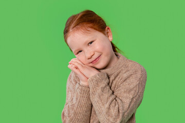 Young girl with red hair smiles sweetly, posing with hands near her face against a green background...