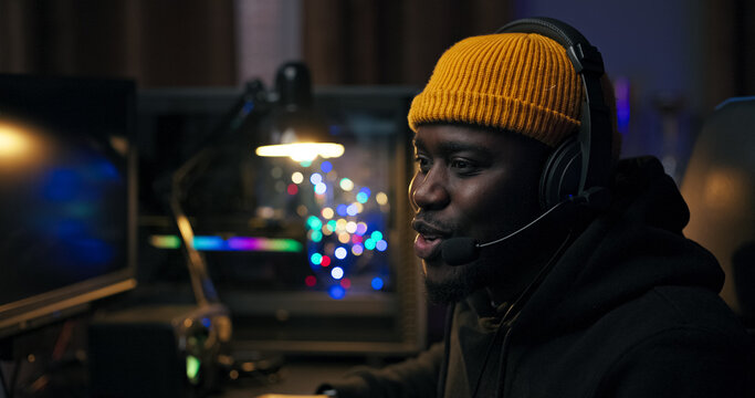 A man sits back in his chair, victorious in an online game. Wearing headphones, he clenches his hands in celebration. The background glows in purple and blue LED light.