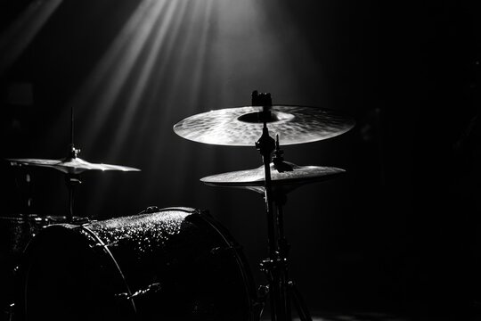 A dramatic black and white close-up of a drum kit under stage lighting, showing detail and texture.
