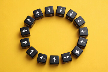 Representing a cycle of choices or paths, black dice with white directional arrows are arranged in a closed circle on a yellow surface, implying movement and decision-making