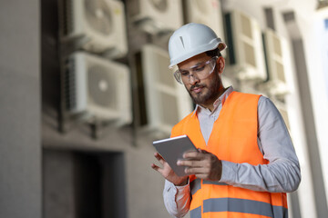 A professional wearing safety gear reviews data on a tablet in front of air conditioning units. The...