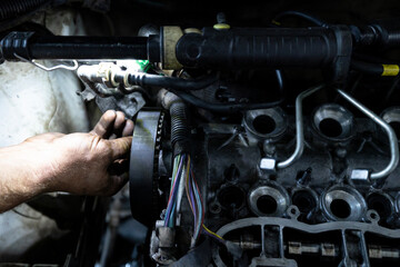 A mechanic's hand works on the timing belt area of a partially disassembled engine inside a vehicle, illuminated by a work light.