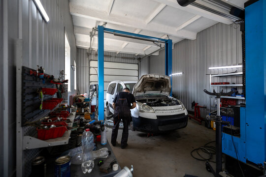 A mechanic works on a white commercial van with its hood open inside a busy auto repair shop equipped with a vehicle lift and various tools
