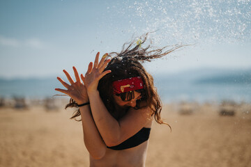 Energetic woman splashes water at a sandy beach, emitting vibrant summer vibes with a playful and happy mood under a clear sunny sky.