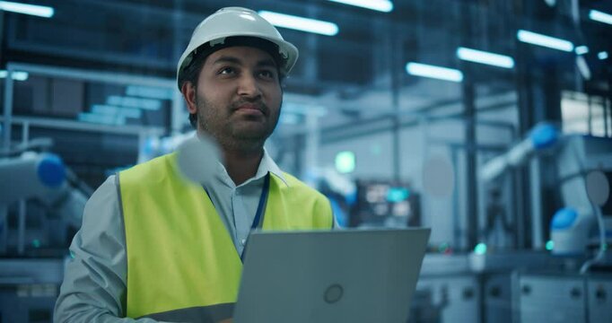 Portrait of an Industrial Systems Administrator Working on Laptop Computer at a Modern Manufacturing Plant. Indian Specialist in a Hard Hat and High Visibility Vest Optimizing Manufacturing Flow