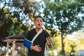 Middle aged Southeast Asian man jogs quickly through park while wearing a medical sling, breathing...