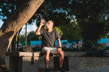 Middle aged Southeast Asian man sits on a bench under shade, rehydrating after a rucking session in...