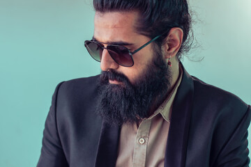 Obraz premium Close-up portrait of man with full, dark beard and long hair tied back in bun. wearing dark sunglasses, beige collared shirt, dark navy blazer. expression is serious thoughtful, gaze directed downward