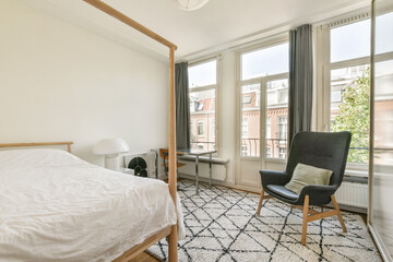 A bright, airy bedroom featuring a wooden bed, modern armchair, and natural light streaming through large windows, creating a serene atmosphere.