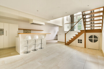 A spacious and bright modern kitchen area featuring sleek design, white cabinetry, bar stools, and a wooden staircase, ensuring a stylish and functional living space.