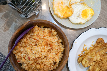 A meal consisting of fried rice served in a wooden bowl with a purple spoon, two fried eggs with runny yolks on a plate, and crispy fried banana on a white scalloped plate, capturing a traditional 
