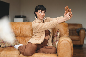 Young woman enjoys a casual selfie moment on a cozy brown sofa in a stylish living room during the...