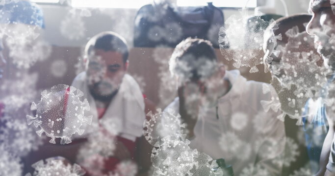 Image of virus cells floating over diverse group of male football players in changing room