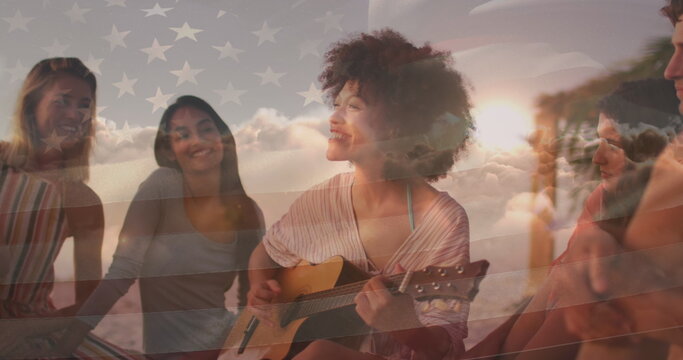 Image of flag of united states of america over happy biracial woman playing guitar on beach - Powered by Adobe