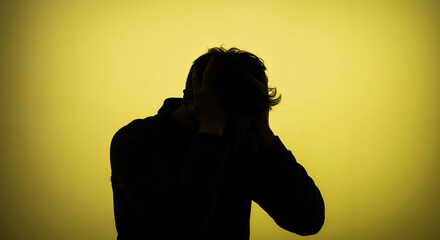 Silhouette of man holding head in hands on yellow background. Person sitting with elbows on knees in distress. Mental health, depression. counseling service, mental health awareness, stress management