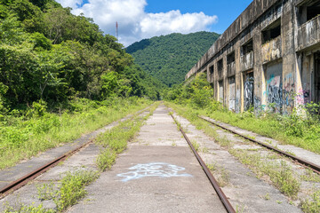 Fototapeta premium Overgrown railway tracks lead to an abandoned factory surrounded by lush mountains in a quiet, chaotic landscape