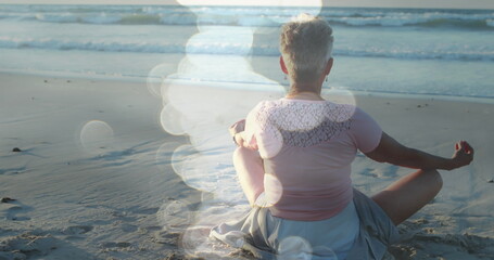 Image of light spots over senior caucasian woman practicing yoga at beach and meditating