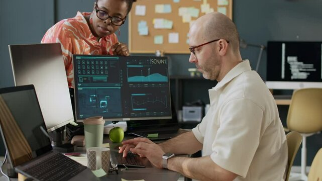 Medium shot of confident young African American female boss checking work of middle-aged Caucasian male programmer showing her new software sitting at computer desk in modern office