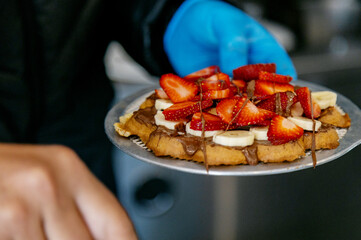 Freshly Made Waffle with Bananas, Strawberries and Chocolate, served on a metal plate. One person is holding the plate, wearing a blue glove