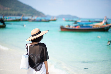 The back of one Asian woman wearing a shirt and a hat with holding bag walks on a sunny beach with blue sea and boats in the background. Concept of holidays and vacations. Picture looking alone or sad
